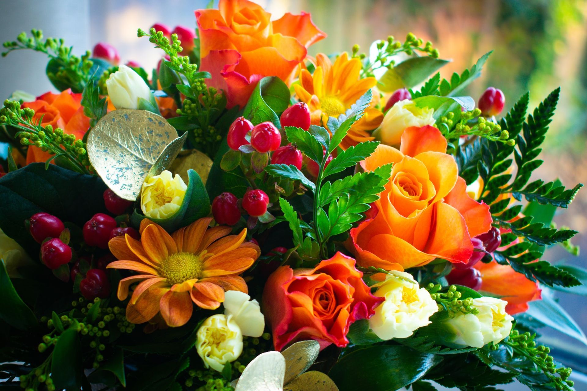 Close-up of a bouquet with orange roses, daisies, berries, and greenery in front of a blurred window.