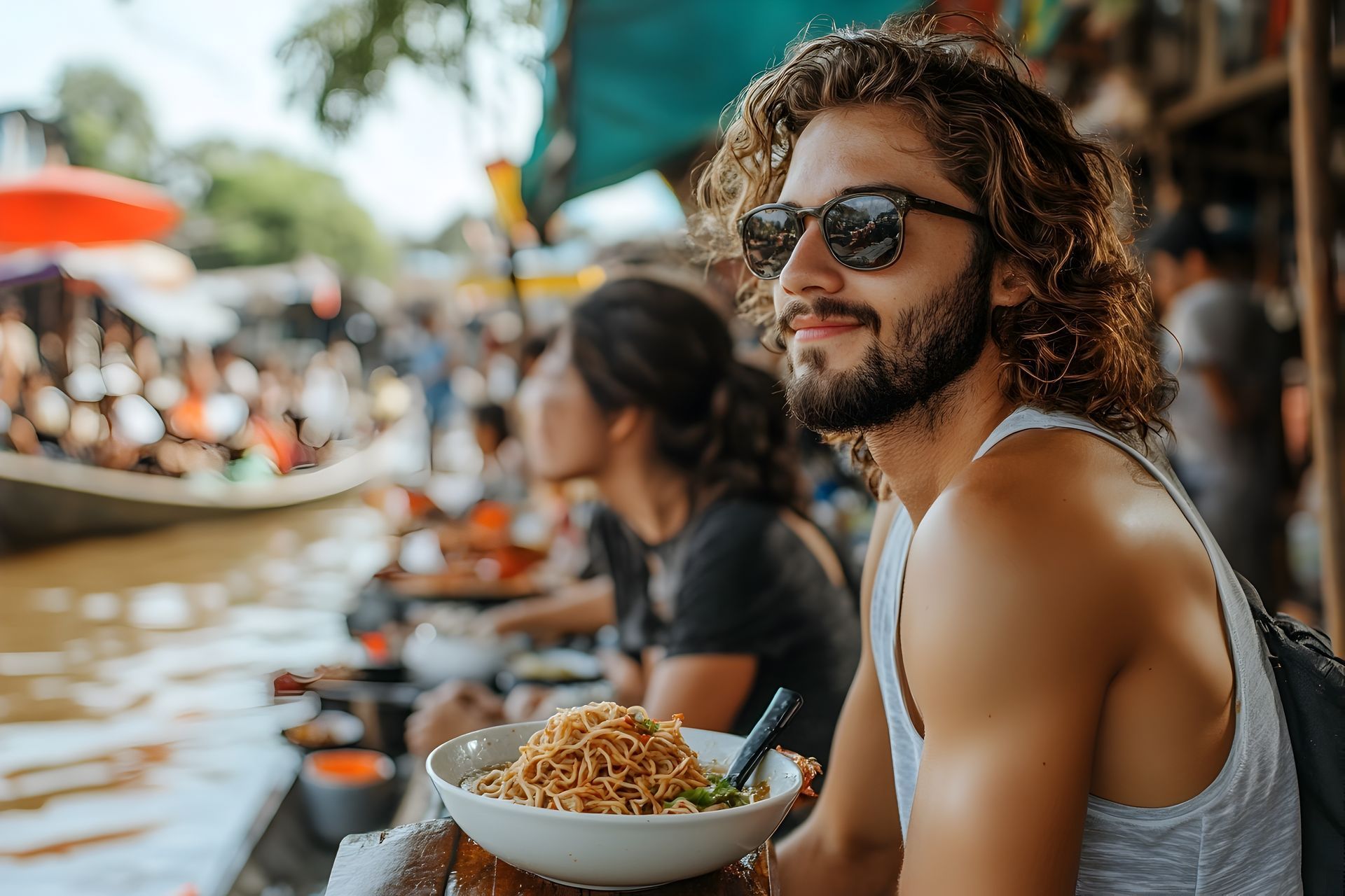 Man with sunglasses and food, sitting at a floating market. Other people are visible in the background.