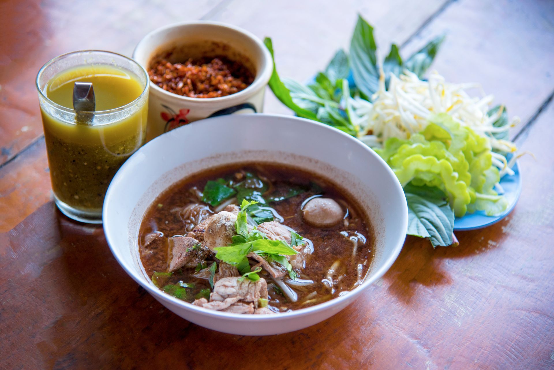 Bowl of dark broth noodles with meat, herbs, and sides of chili, vegetables, and a drink.