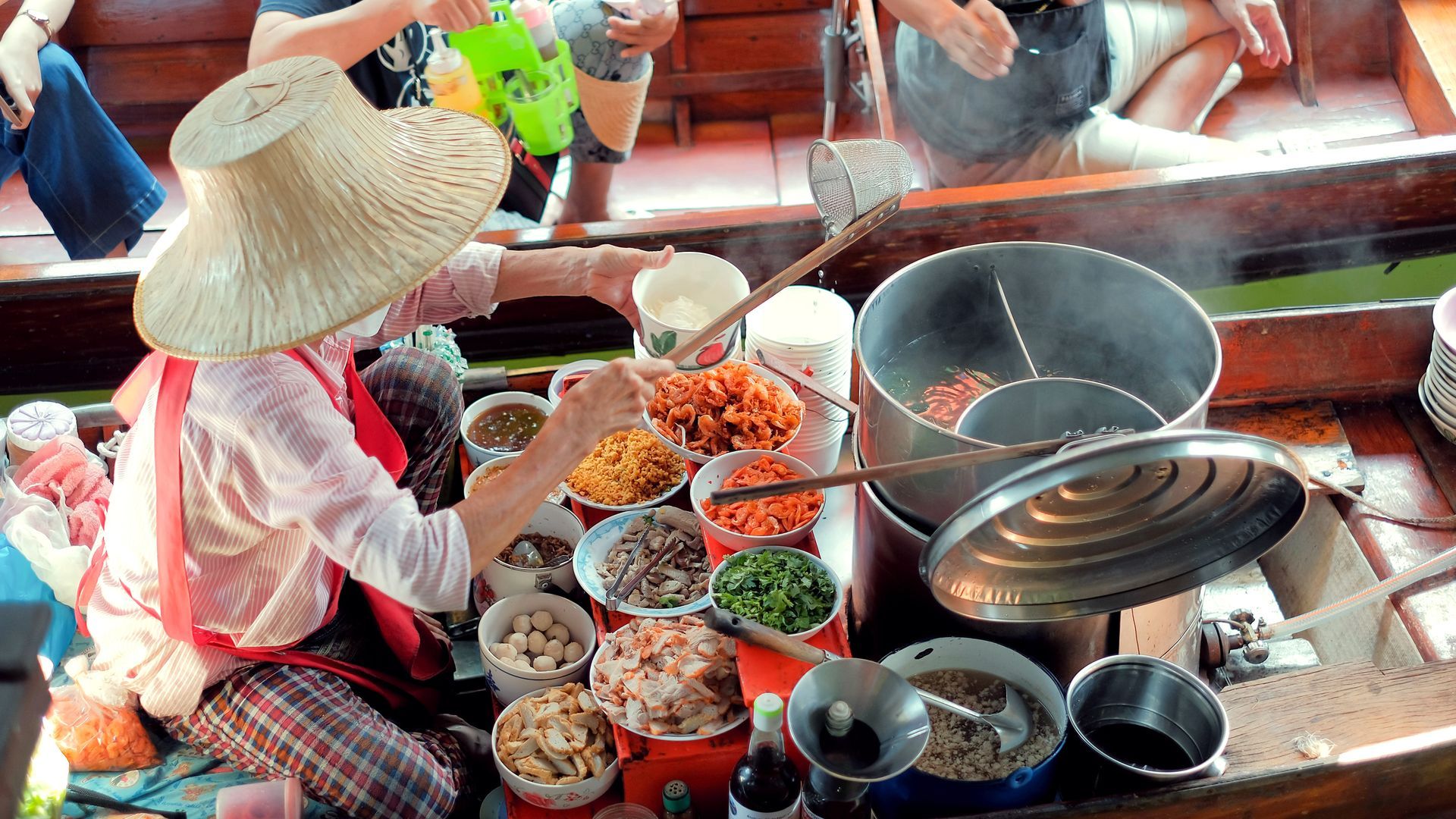 Woman in straw hat prepares food on a boat, various ingredients are in bowls, customers are in the background.
