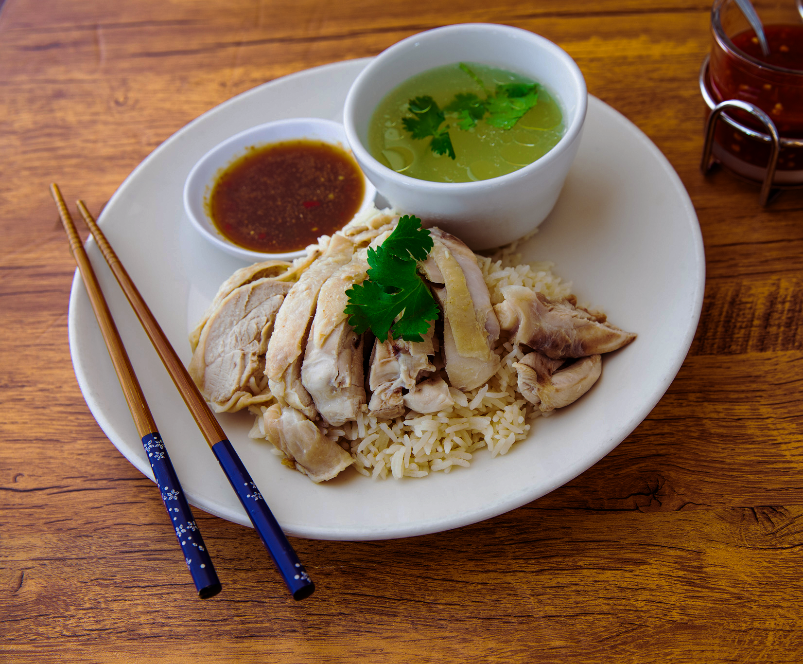 Hainanese chicken rice: sliced chicken over rice, broth, sauce, chopsticks, small dish of chili sauce on a white plate.