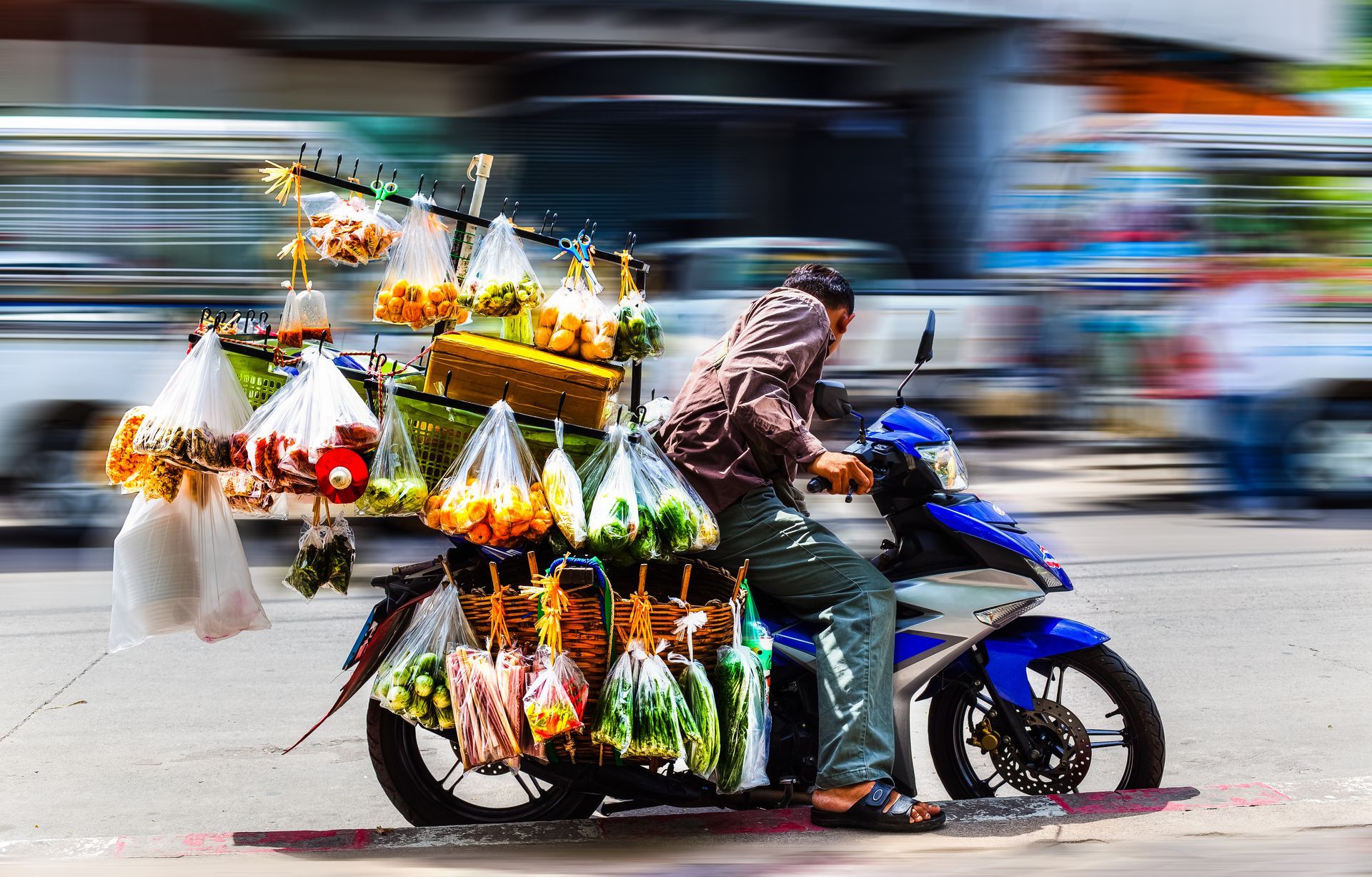 Man rides a blue motorcycle overloaded with bags of fruit, likely selling them.