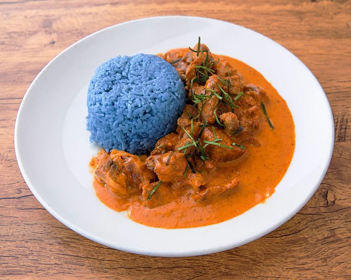 Plate of red curry, beef stew with blue rice on a wooden surface.