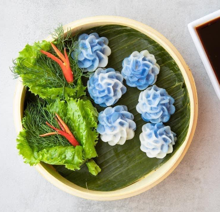 Blue flower-shaped Thai dumplings on a plate with herbs and dipping sauce.