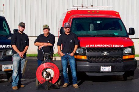Three workers stand by service vans, one with a cable cleaning machine. Vans are red and black.