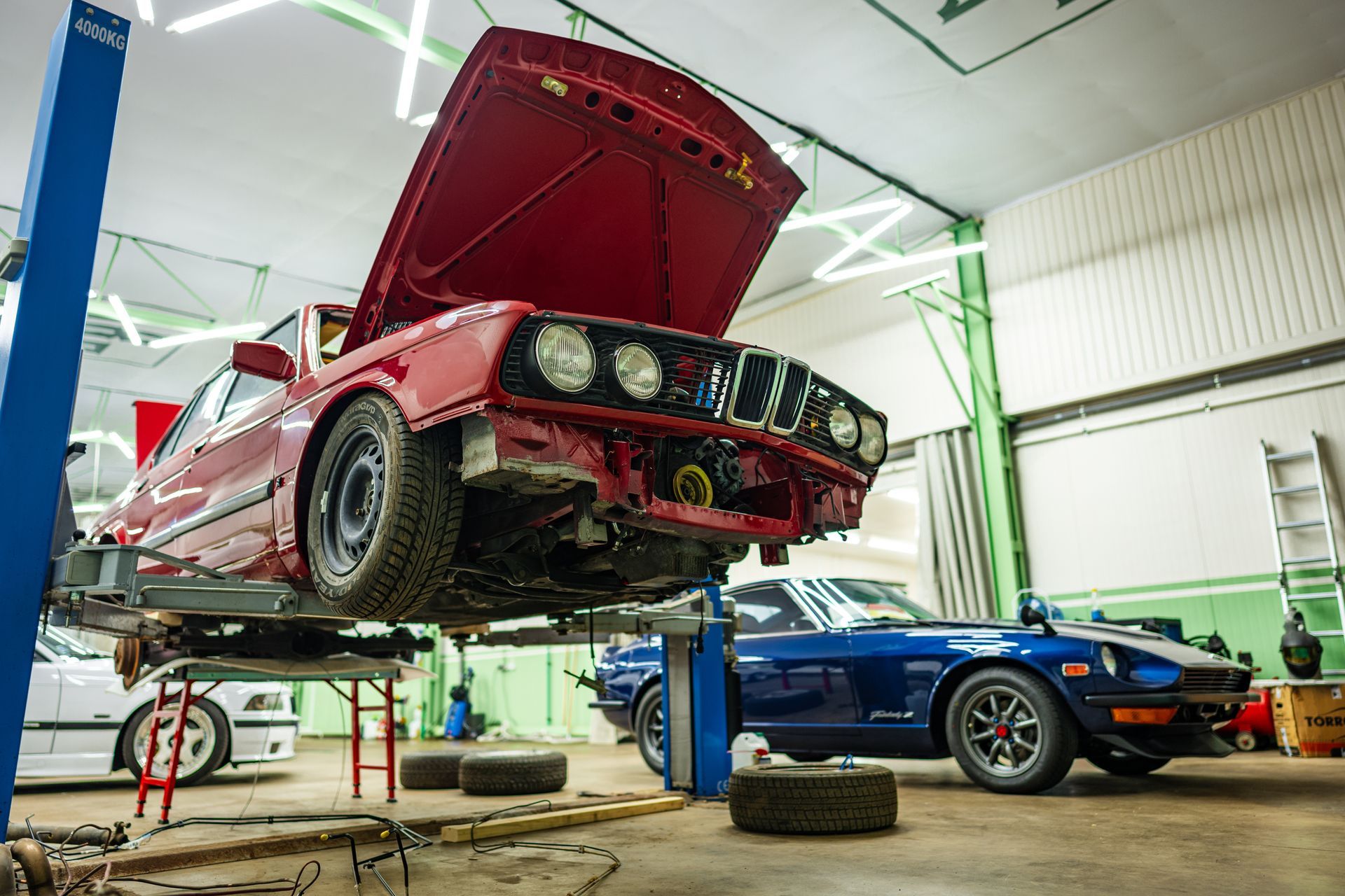 Red car on a lift in a garage, hood open. Blue car and another vehicle visible.