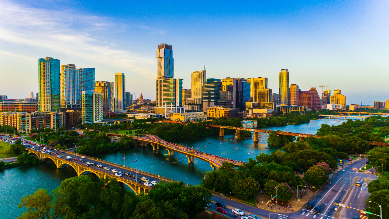 Aerial view of a modern city skyline beside a river, with bridges, roads, and trees at sunset.