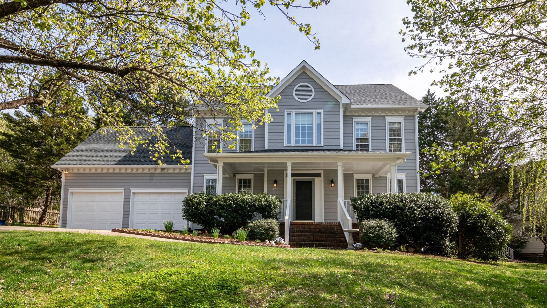 A two-story, gray house with a front porch, an attached garage, and a lush front lawn surrounded by mature trees.