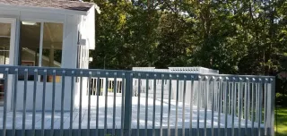 White house deck with blue railing in a wooded yard, with a door and windows visible.