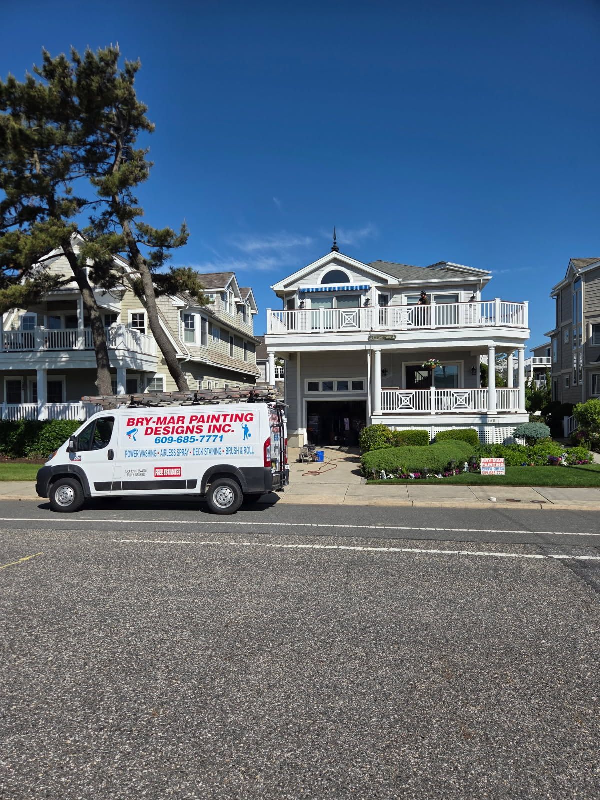 White two-story house with a blue roof and a bry-mar painting & designs van parked in front on a sunny street.
