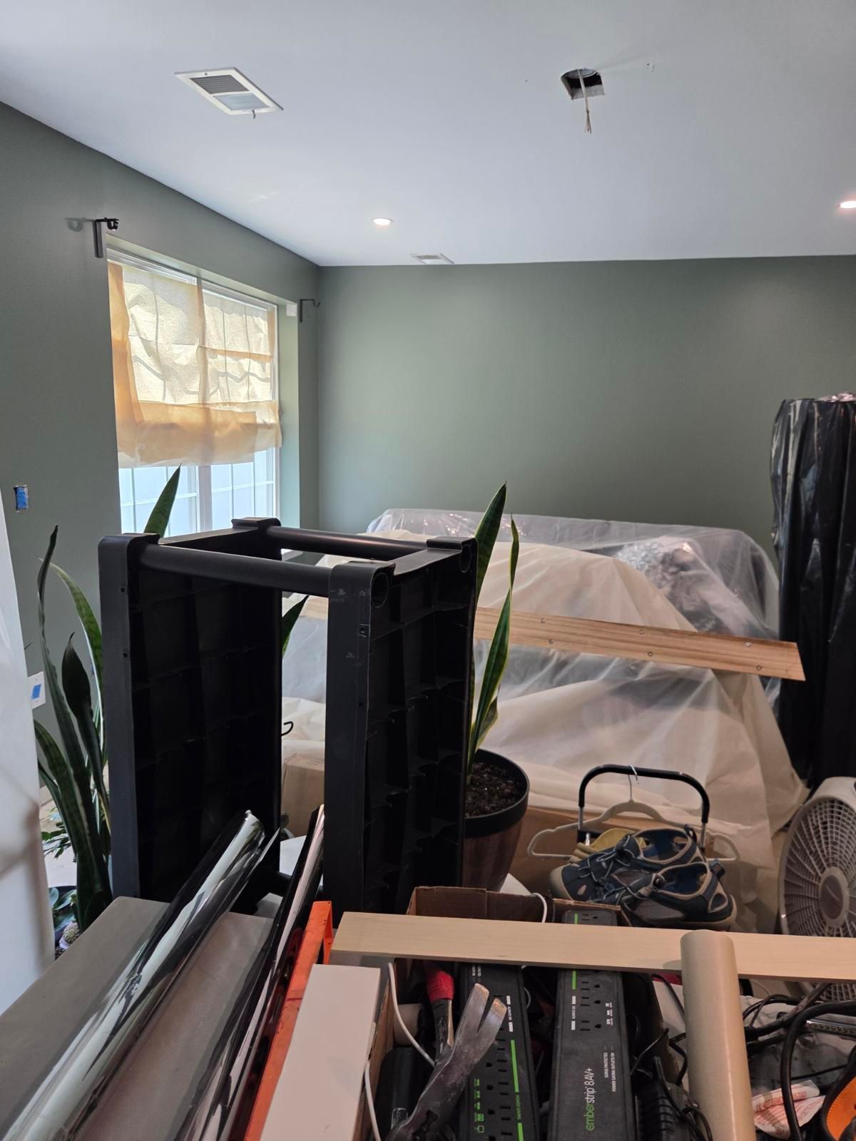 Cluttered bedroom with furniture parts, plants, and a bed against gray walls and a window.