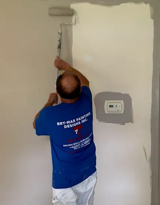 Painter rolling white paint on a wall beside a thermostat in a room