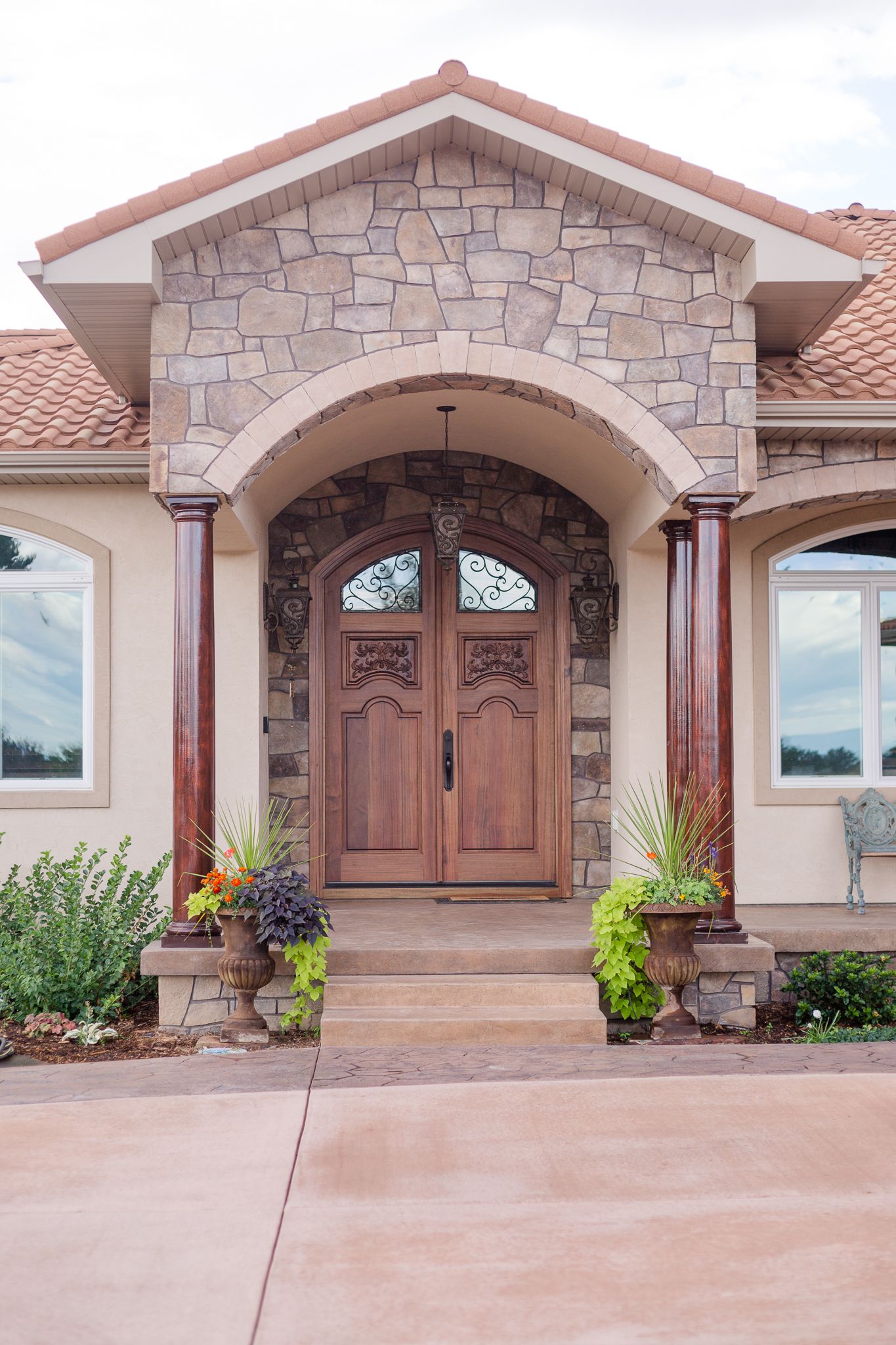 Front entrance of a beige home with stone archway, brown doors, and flowerpots.