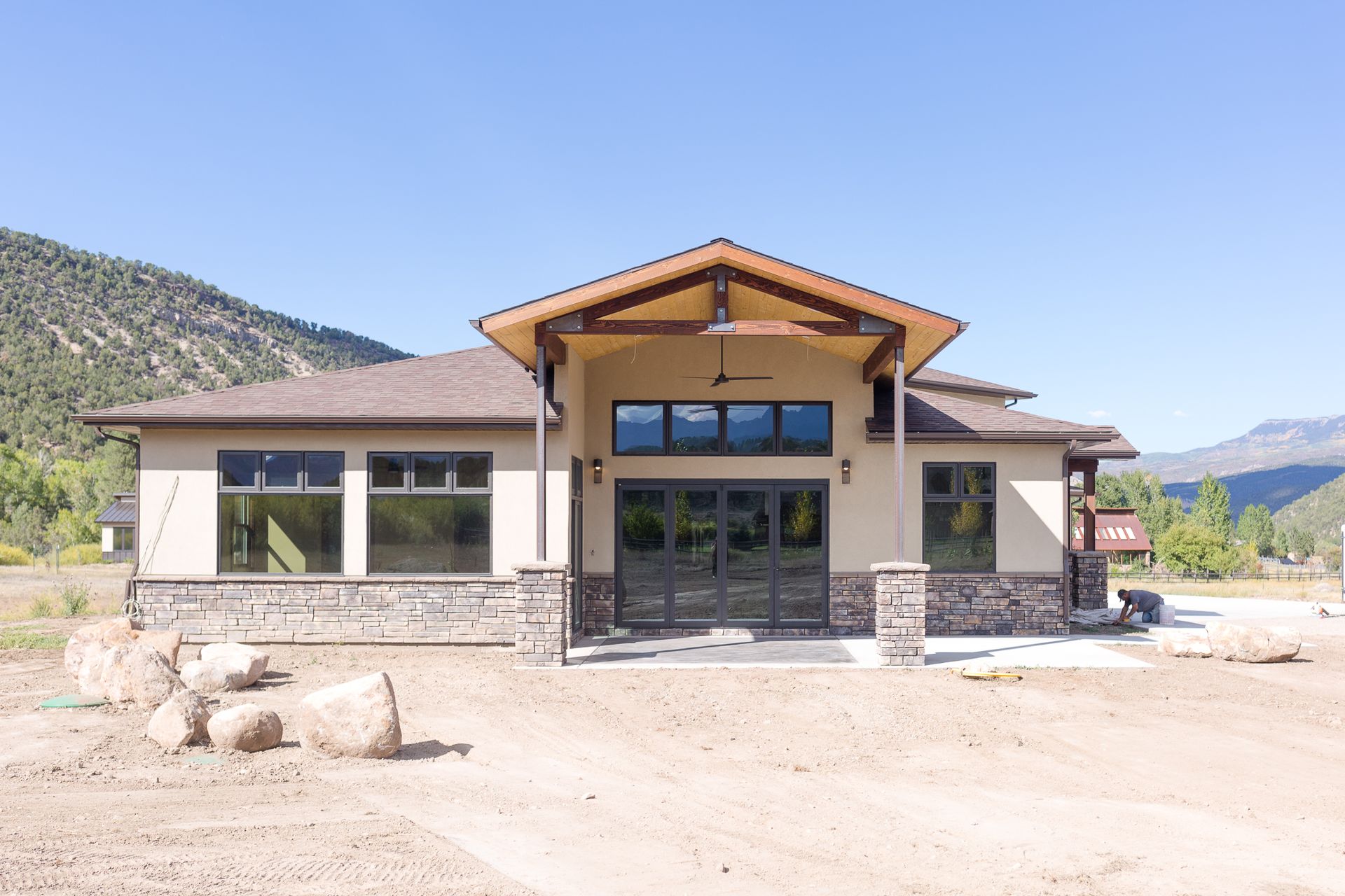 Tan building with large windows, stone accents, and a wooden beam entrance, set against a mountain backdrop.