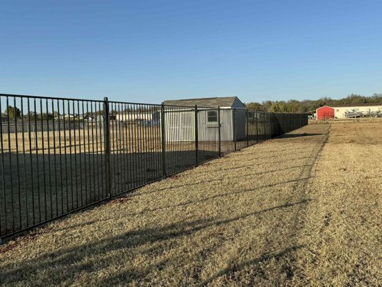 Black metal fence beside a gravel yard with a white shed and red structure under a clear blue sky