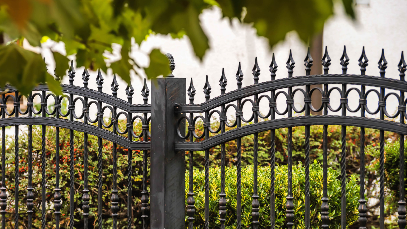 Black wrought-iron fence with pointed finials in front of green shrubs and trees