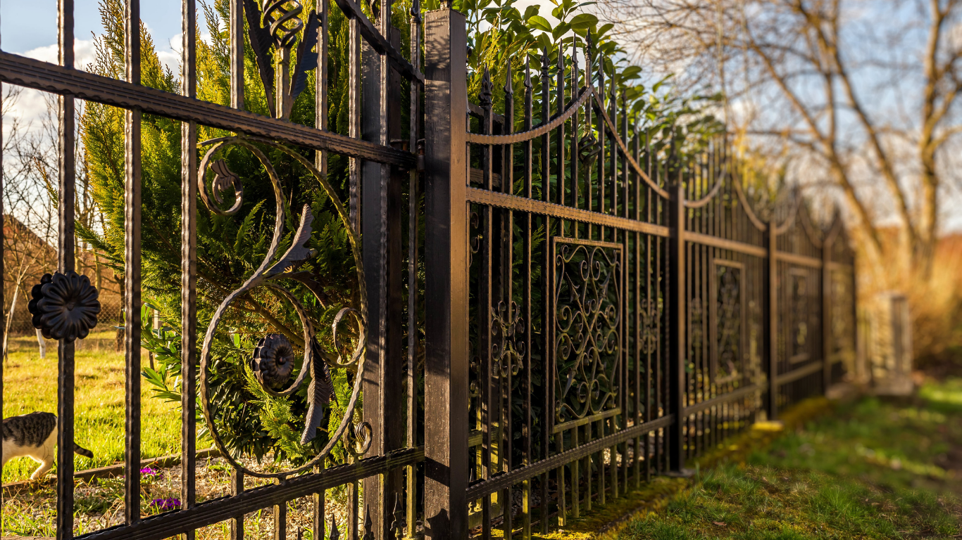 Black wrought-iron fence along a sunlit path with trees and greenery behind it