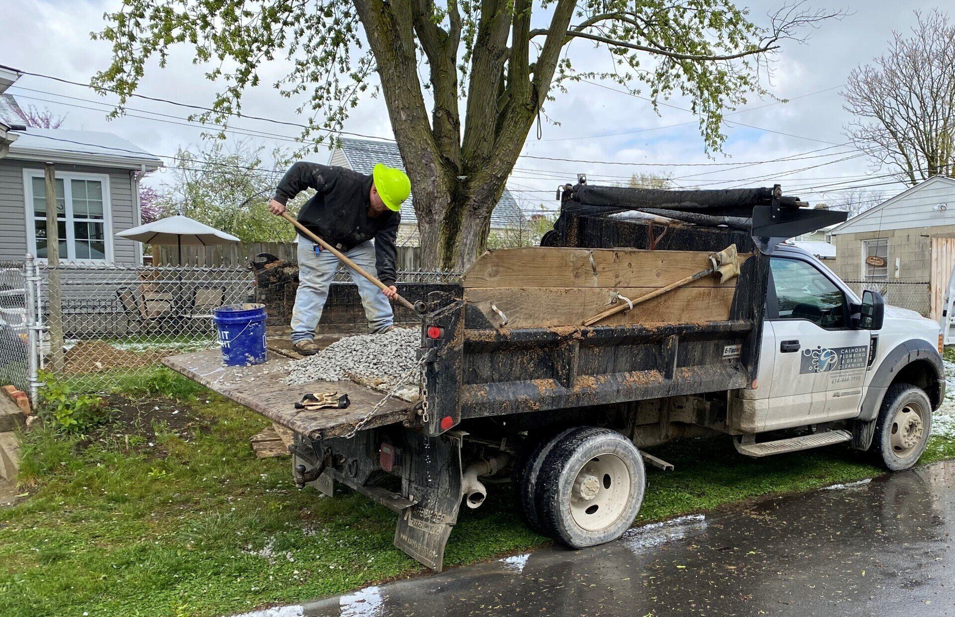 Man shoveling gravel from a truck bed near a tree in a residential area.