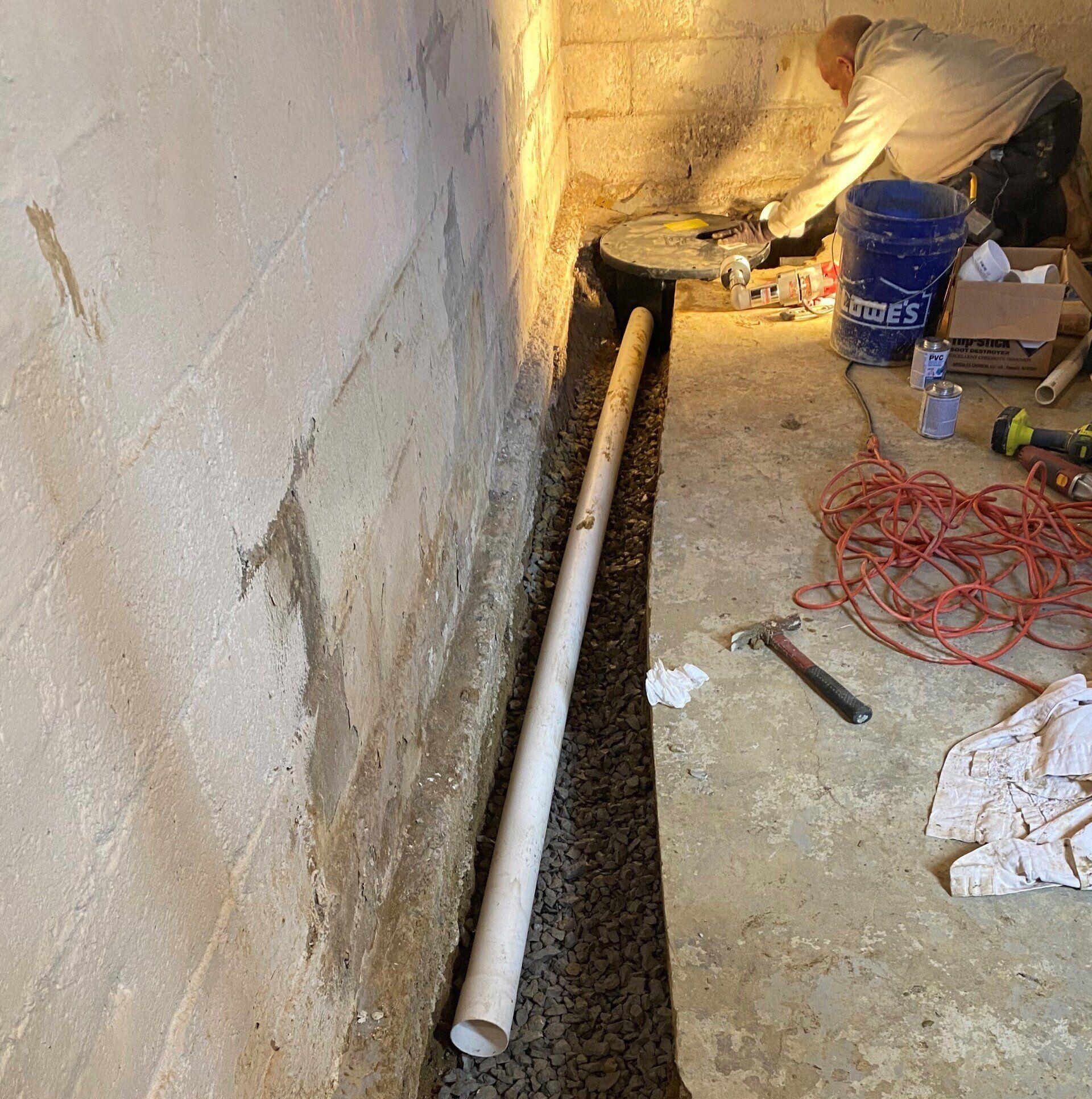 A person working on a basement drainage system. White pipe and gray gravel fill a trench near a concrete wall.