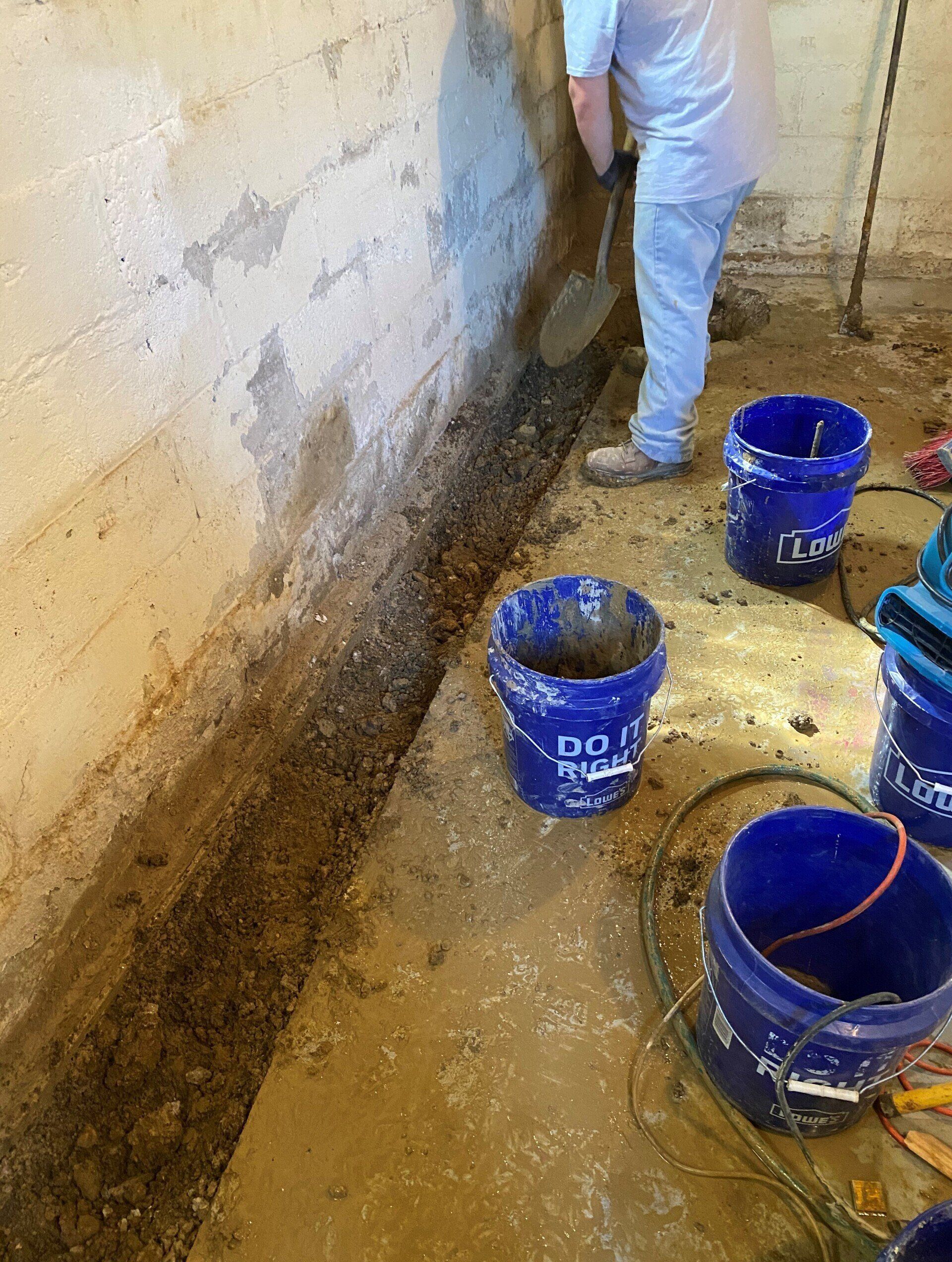 Person shoveling gravel from a trench next to a concrete basement wall; three blue buckets nearby.