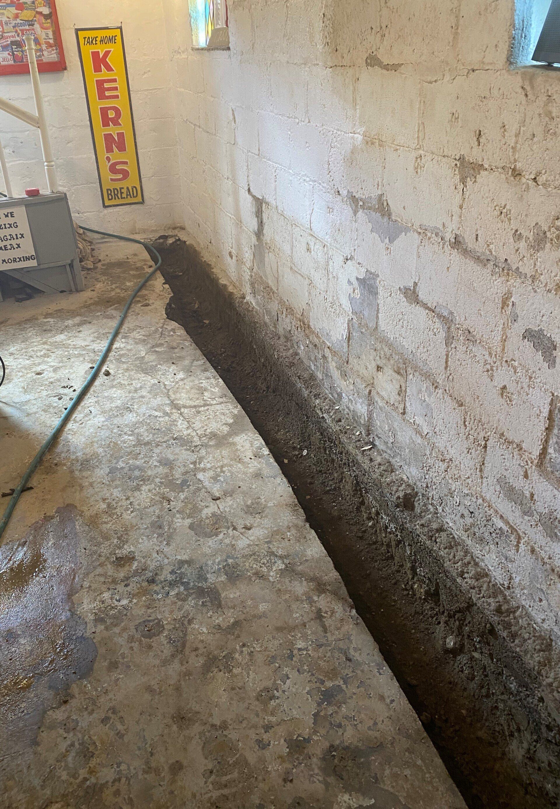 Long trench dug along a basement wall, filled with dirt and debris.  A hose lies nearby.