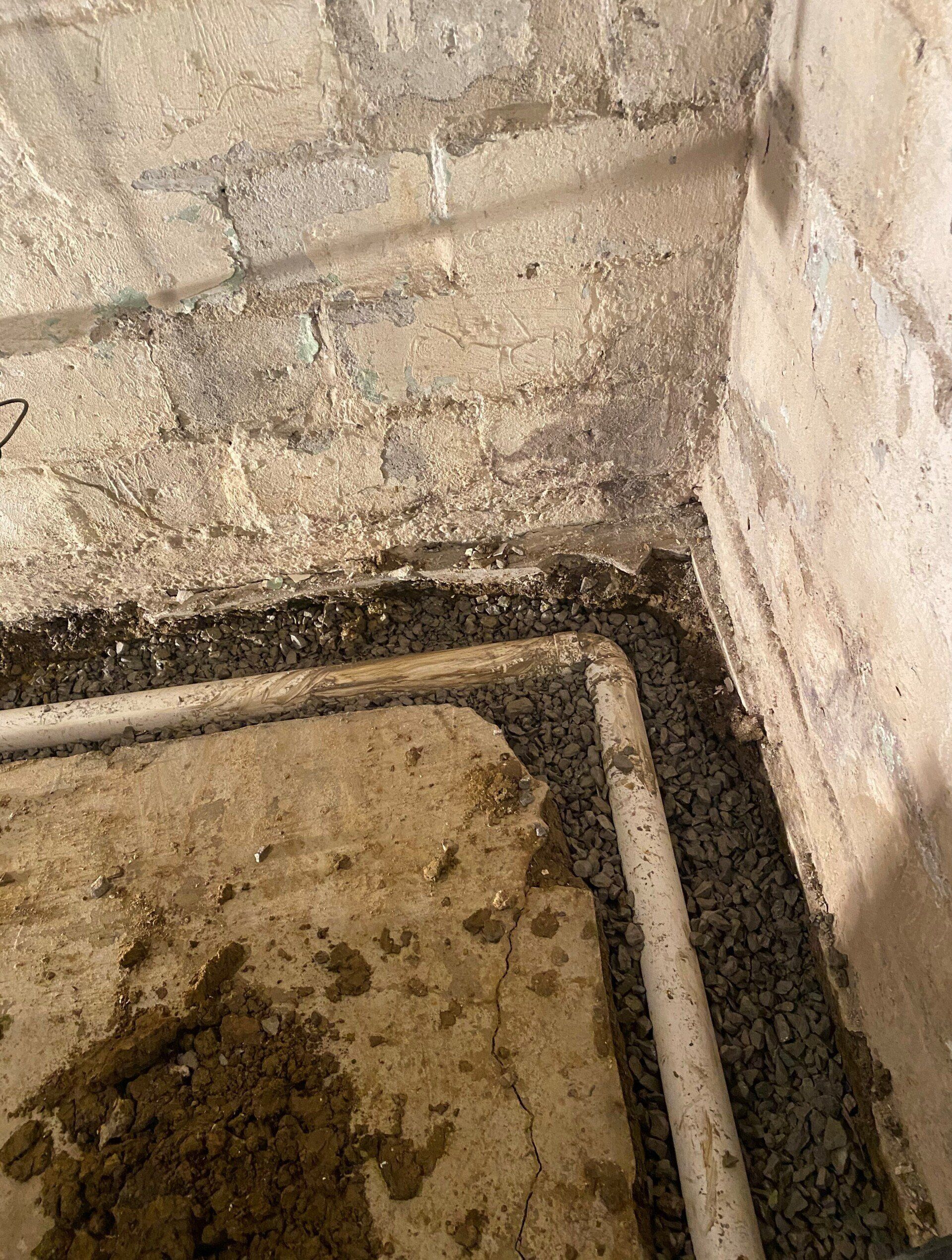 Concrete basement corner with exposed piping and debris, likely showing construction or damage.