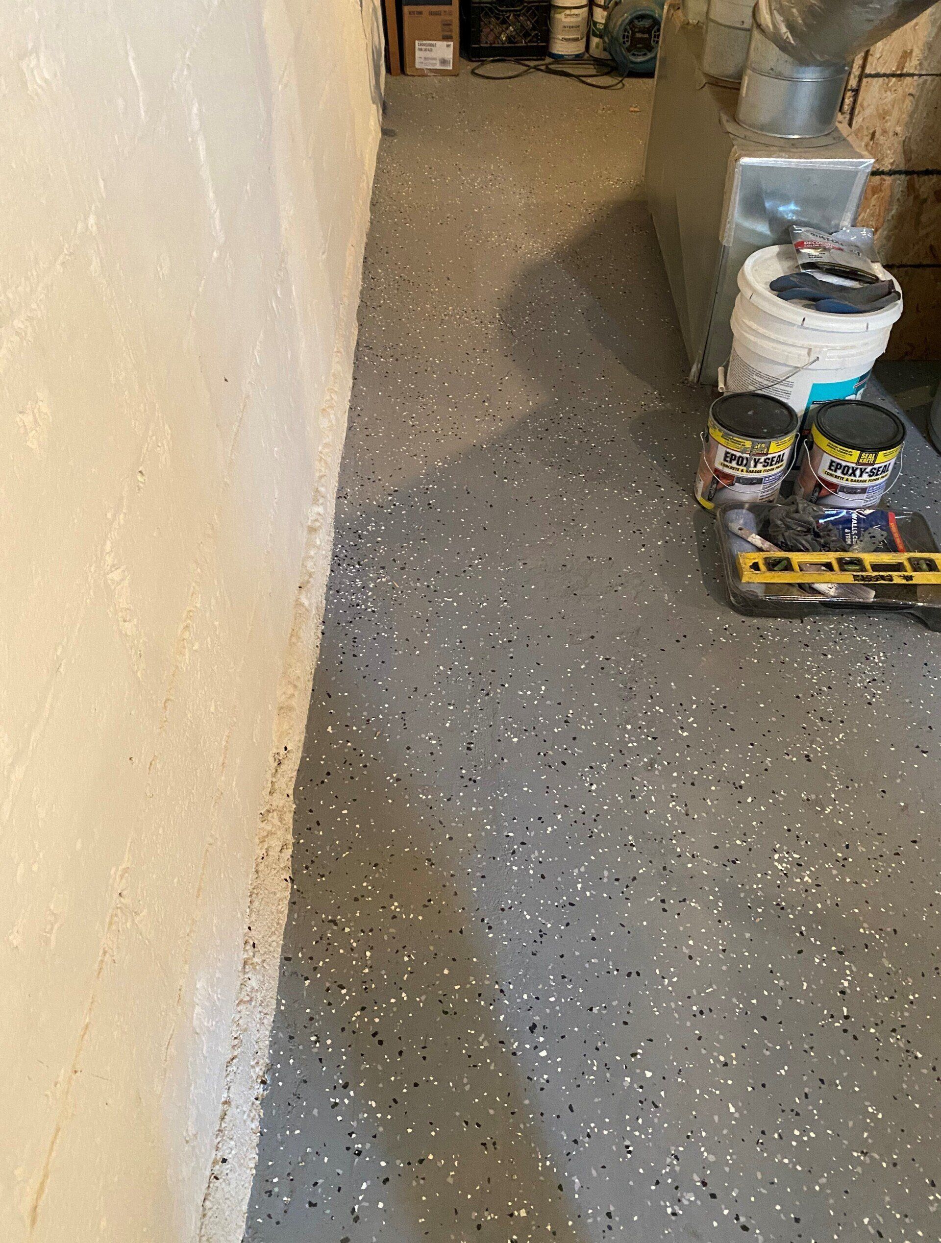 Concrete floor with speckled pattern along a basement wall, with paint cans and equipment nearby.