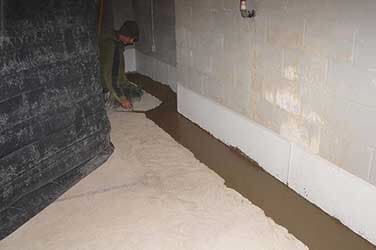 Person working in a basement, applying sealant along a wall, with a layer of waterproof membrane.