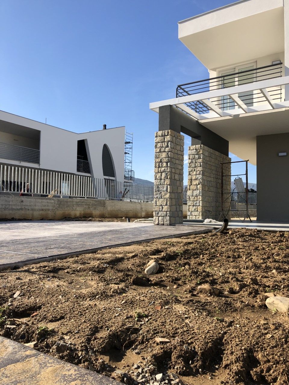 Esterno di una casa moderna con colonne in pietra, balcone e un primo piano di terra nuda sotto un cielo azzurro.