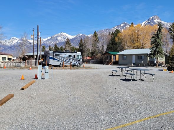A graveled RV park with picnic tables in the foreground, featuring a parked RV and mountains under a clear blue sky.