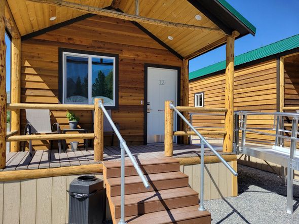 A wood cabin with a covered front porch, stairs with metal railings, and a white door, set in an outdoor area.