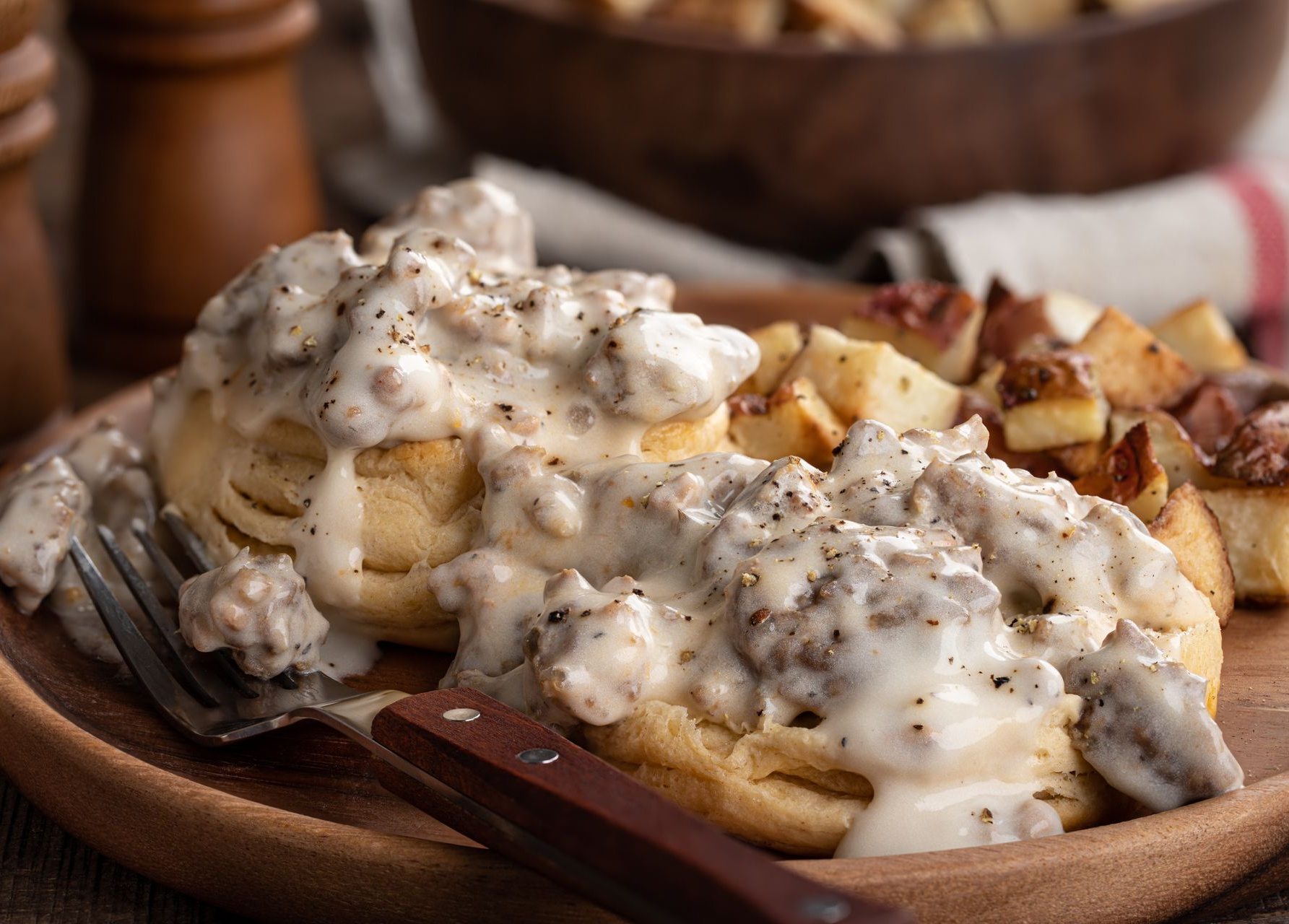 Biscuits and sausage gravy on a wooden plate, accompanied by roasted breakfast potatoes.