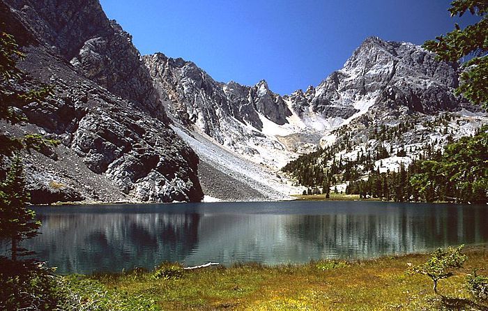 A serene, clear mountain lake reflecting craggy, snow-dusted peaks under a bright, cloudless blue sky.