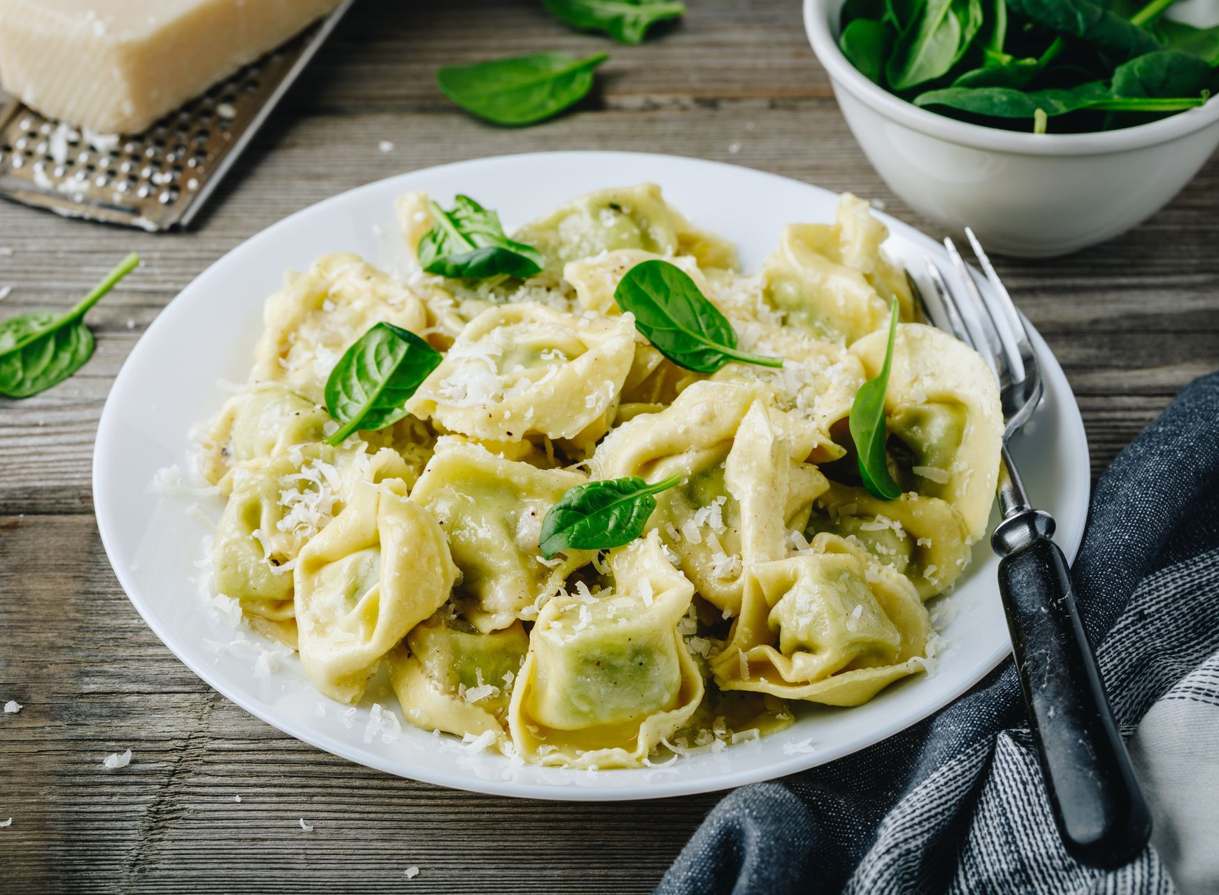 A white plate of tortellini sprinkled with parmesan and topped with fresh spinach leaves, set on a rustic wooden table.
