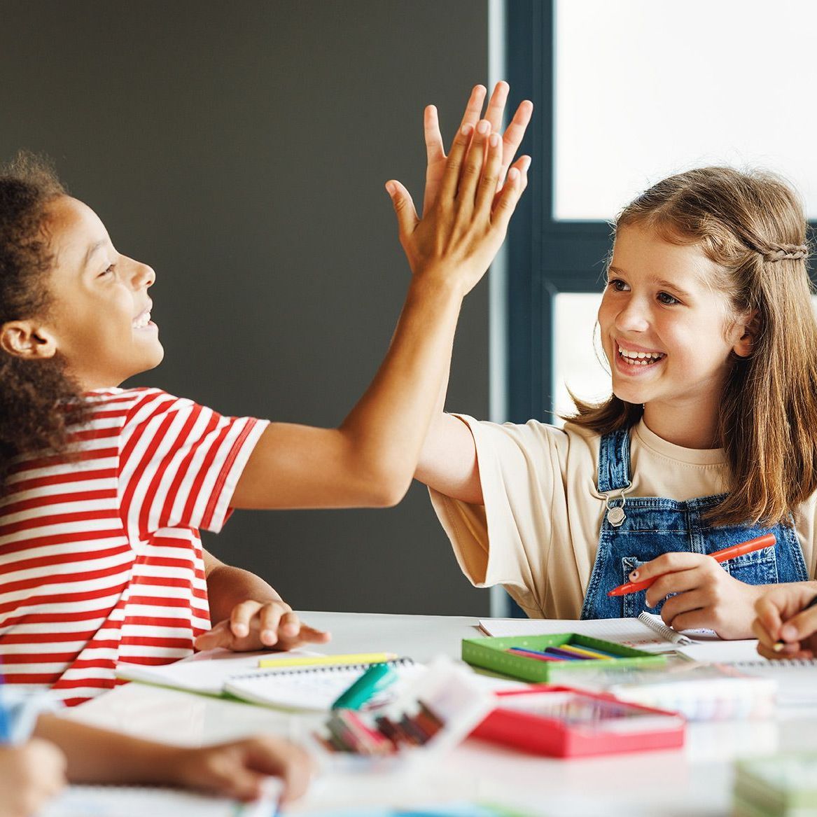 Two young girls are giving each other a high five while sitting at a table.