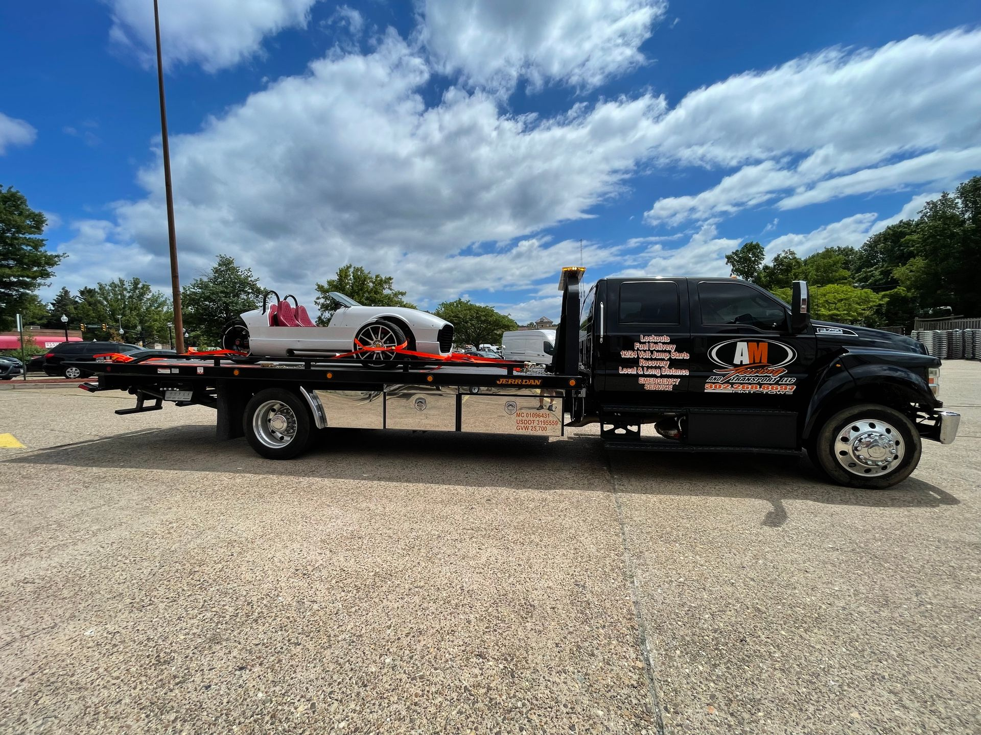 Black tow truck with white car on flatbed, parked on gravel against a cloudy sky.
