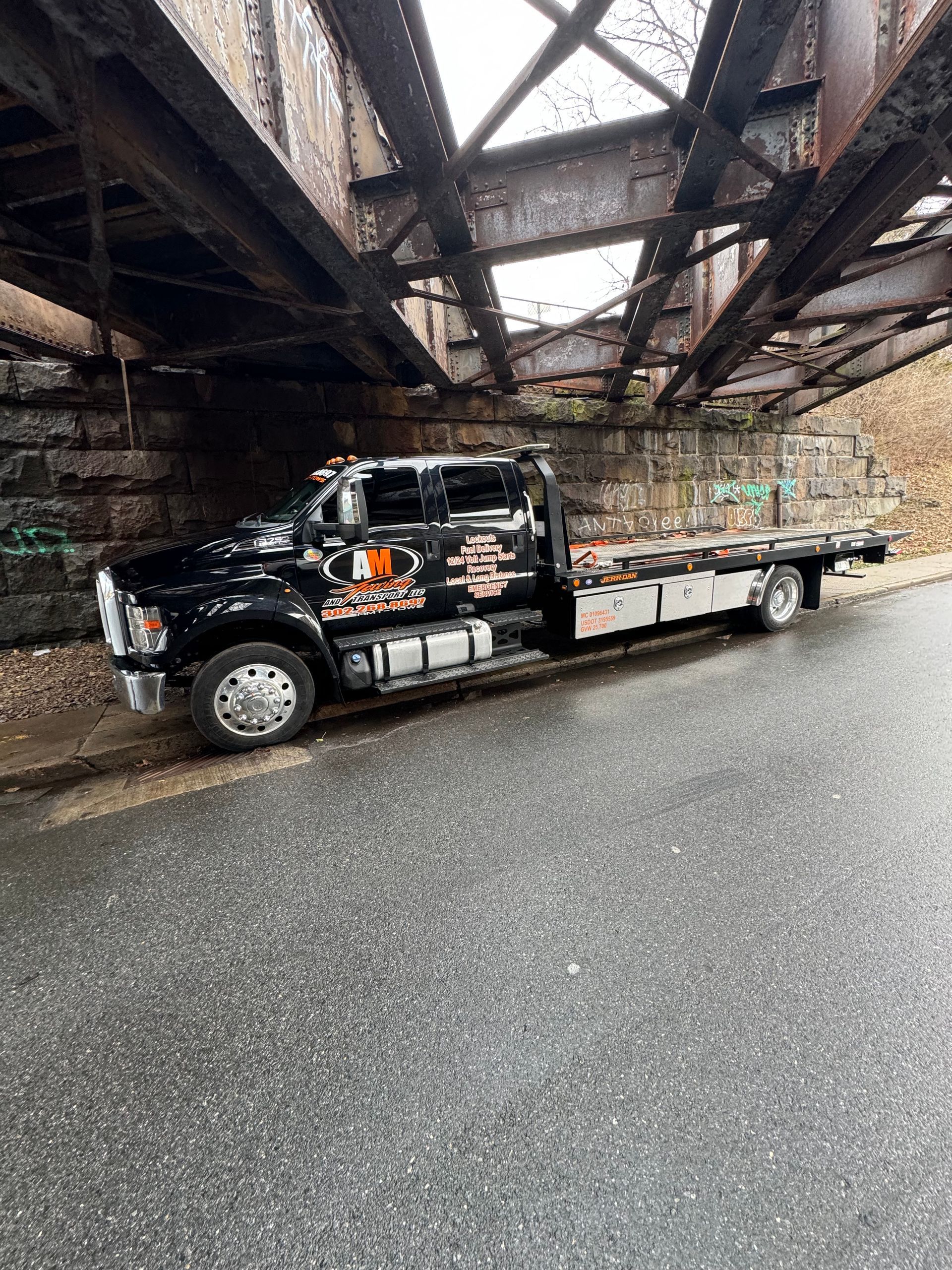 Black tow truck parked under a weathered, metal bridge. Overcast day.
