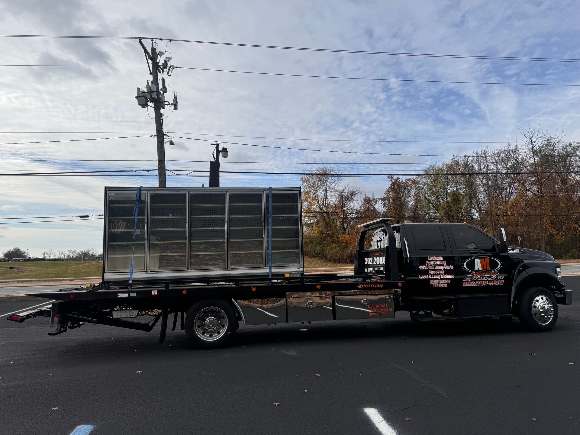 Black tow truck carrying a large metal structure, parked on asphalt. Overcast sky.