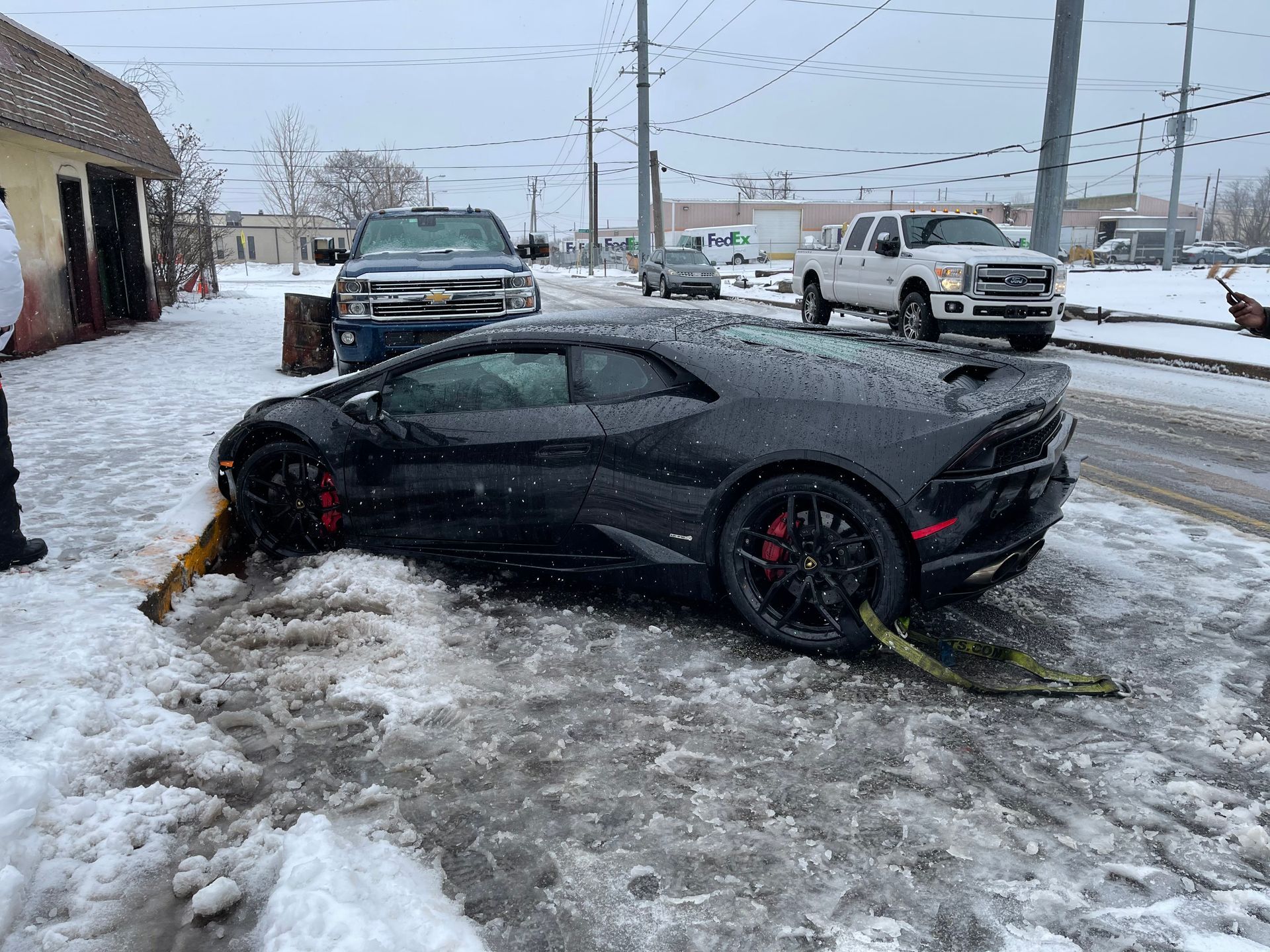 Black Lamborghini stuck in snow on a city street; tow straps attached.
