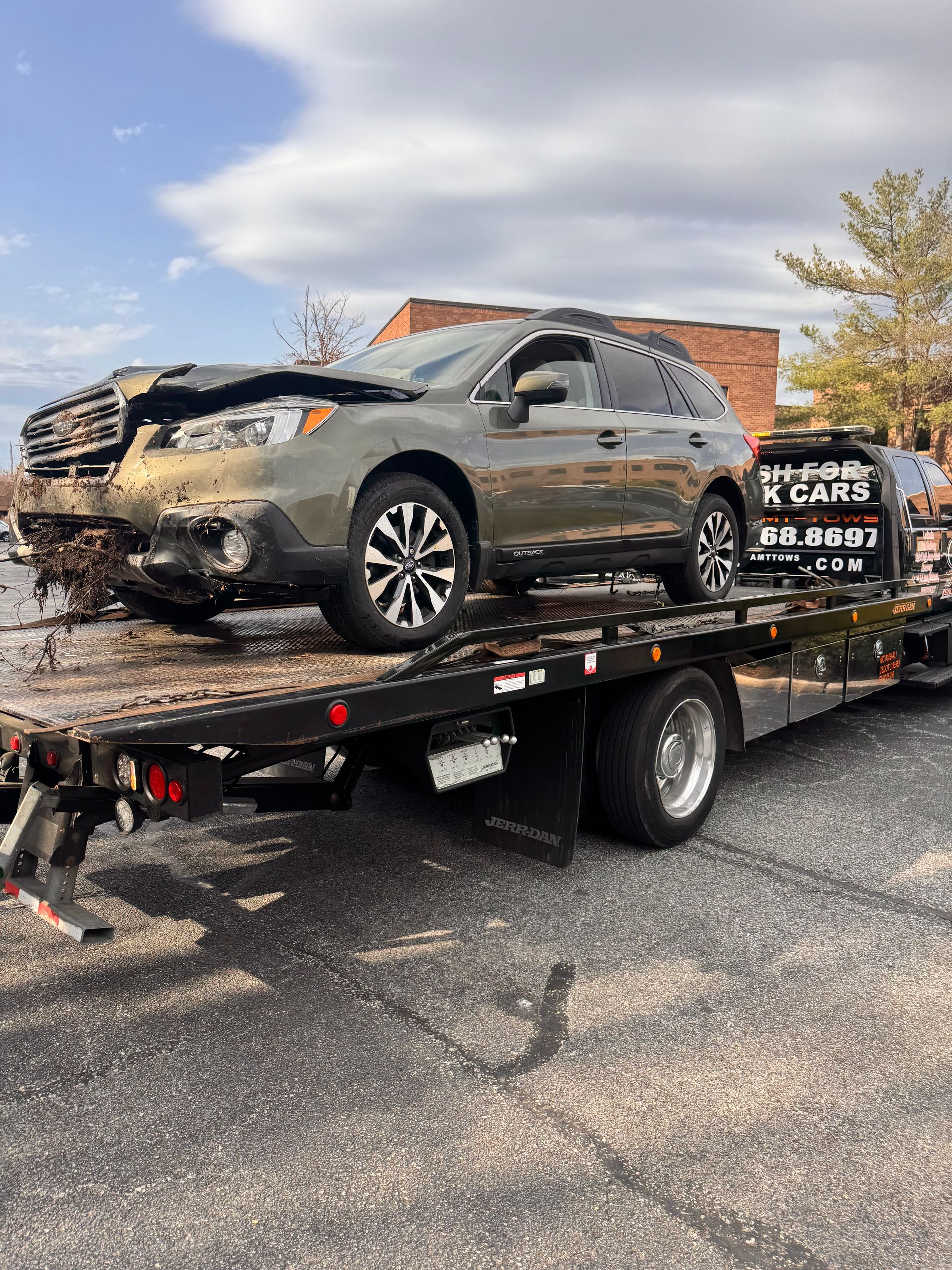 Damaged green Subaru Outback on a tow truck, front end heavily damaged, on a paved surface near a building.