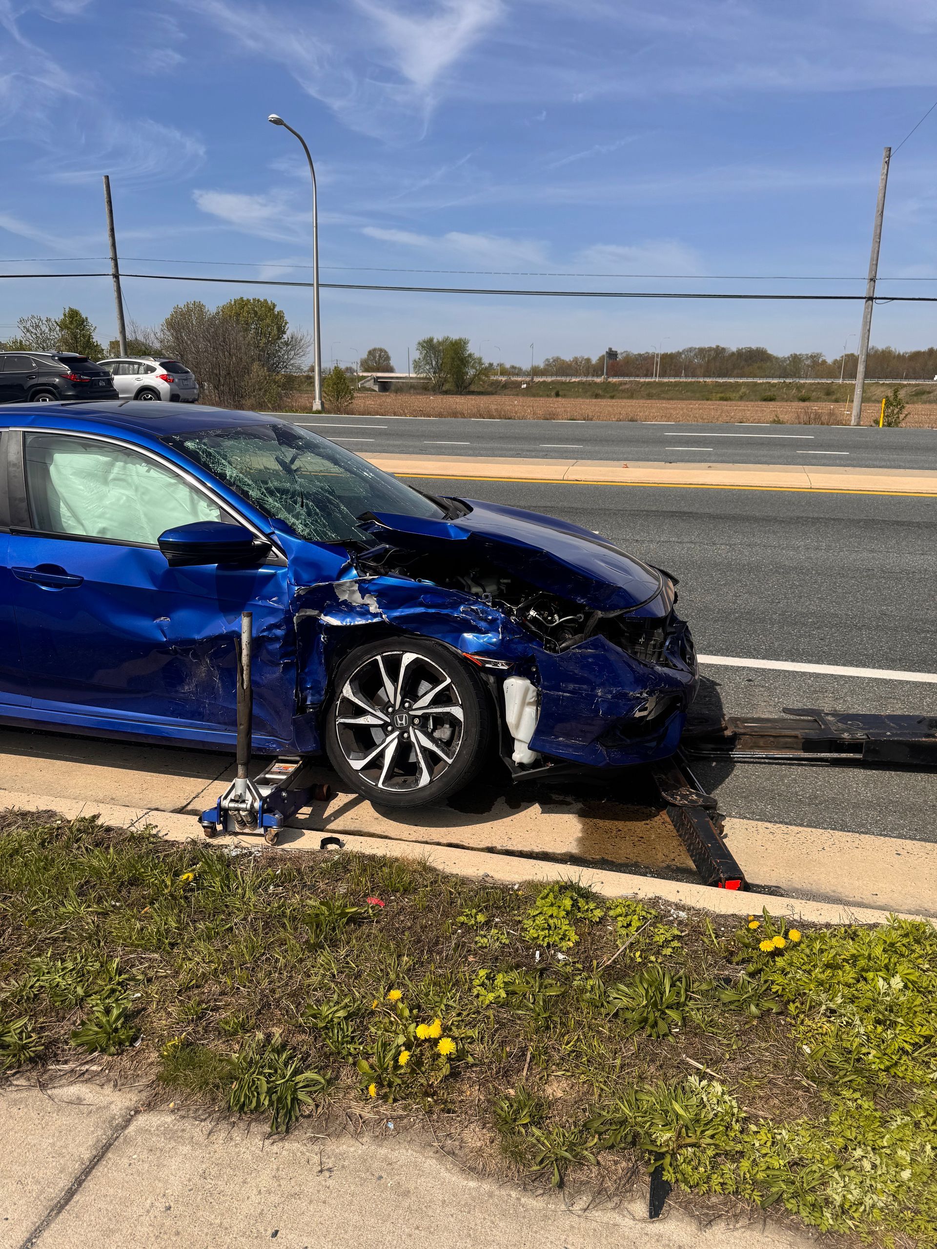 Blue car with significant front-end damage, airbag deployed, roadside setting with grass and asphalt.