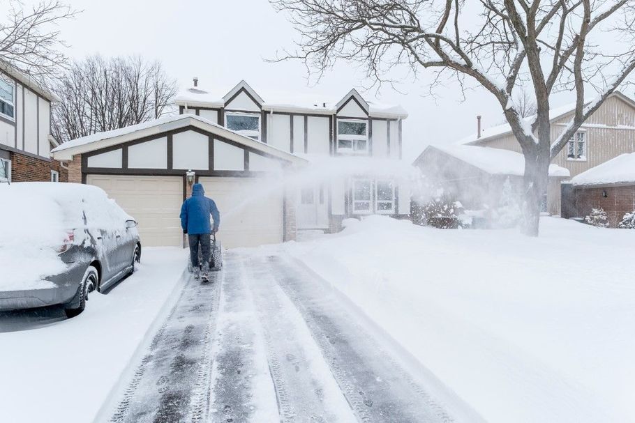 Snow-covered suburban driveway with a person shoveling snow beside parked cars and houses.