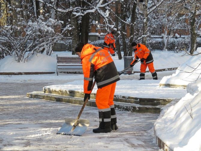 Workers in bright orange gear shoveling snow from a frozen path in a snowy park