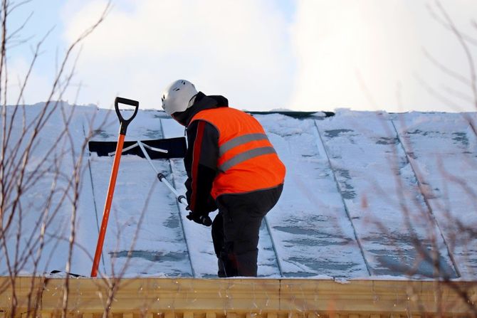 Worker in orange safety vest clearing snow from a rooftop with a shovel