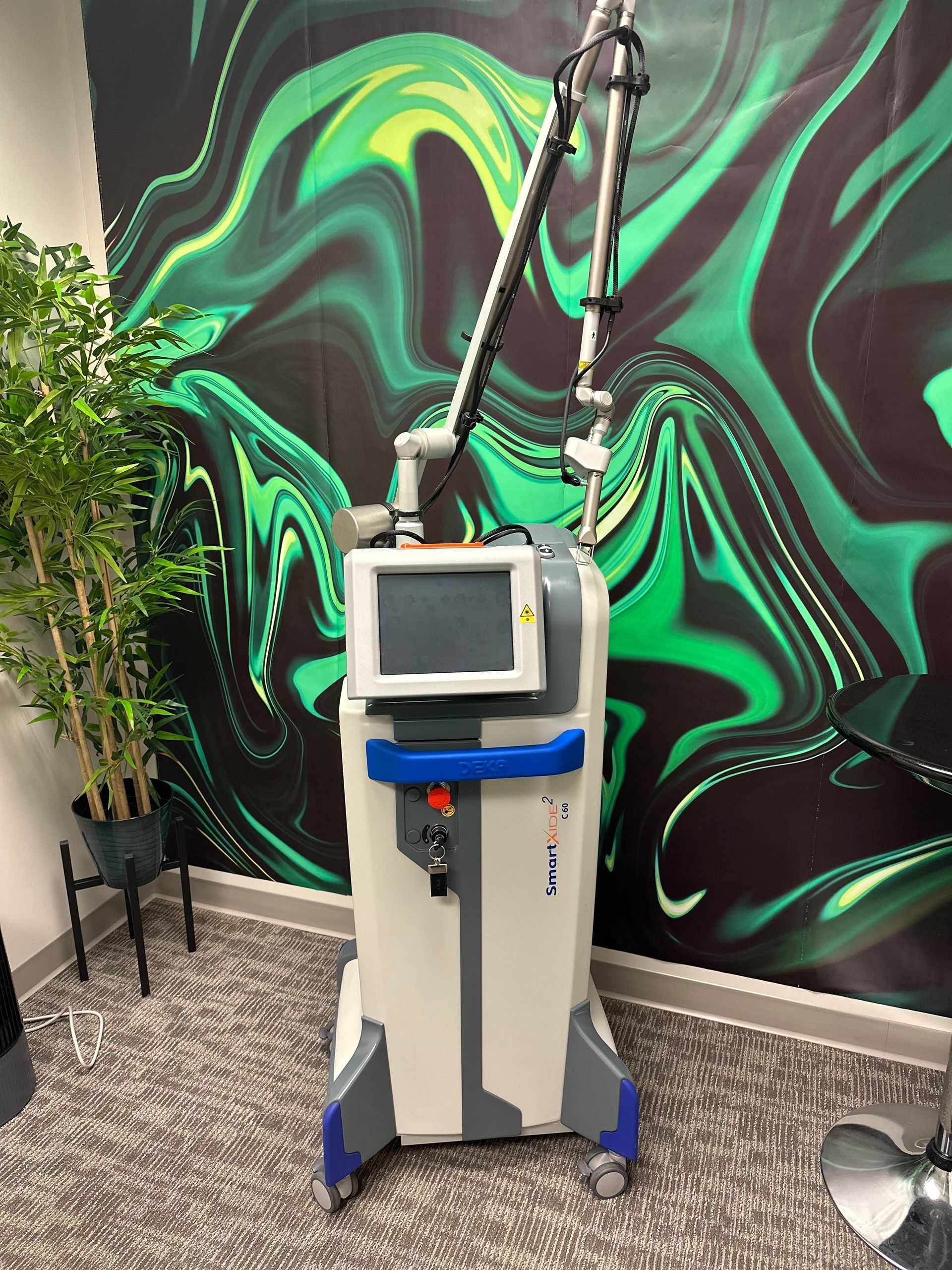 Laser machine in a room, with a digital display and articulated arm, against a green and black abstract wall.