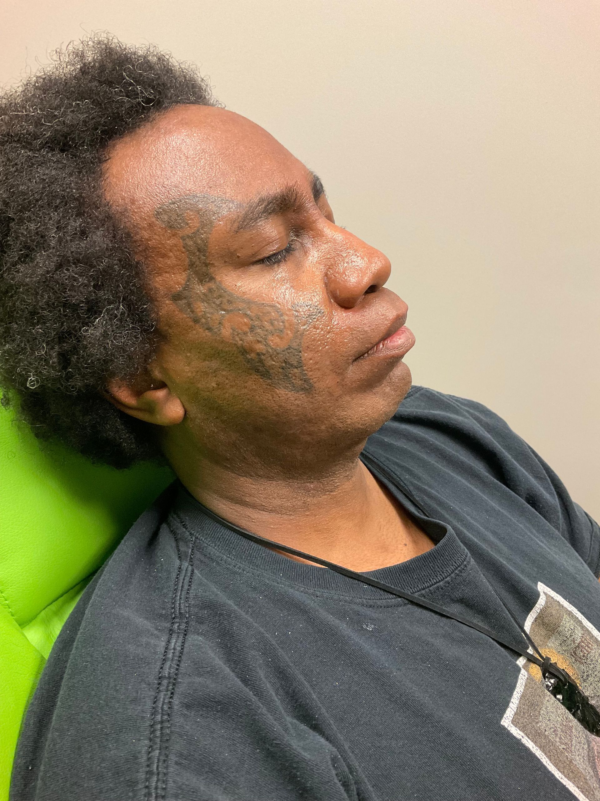 Man with dark skin, eyes closed, resting in a chair. Patchy facial hair. Wearing a dark t-shirt.
