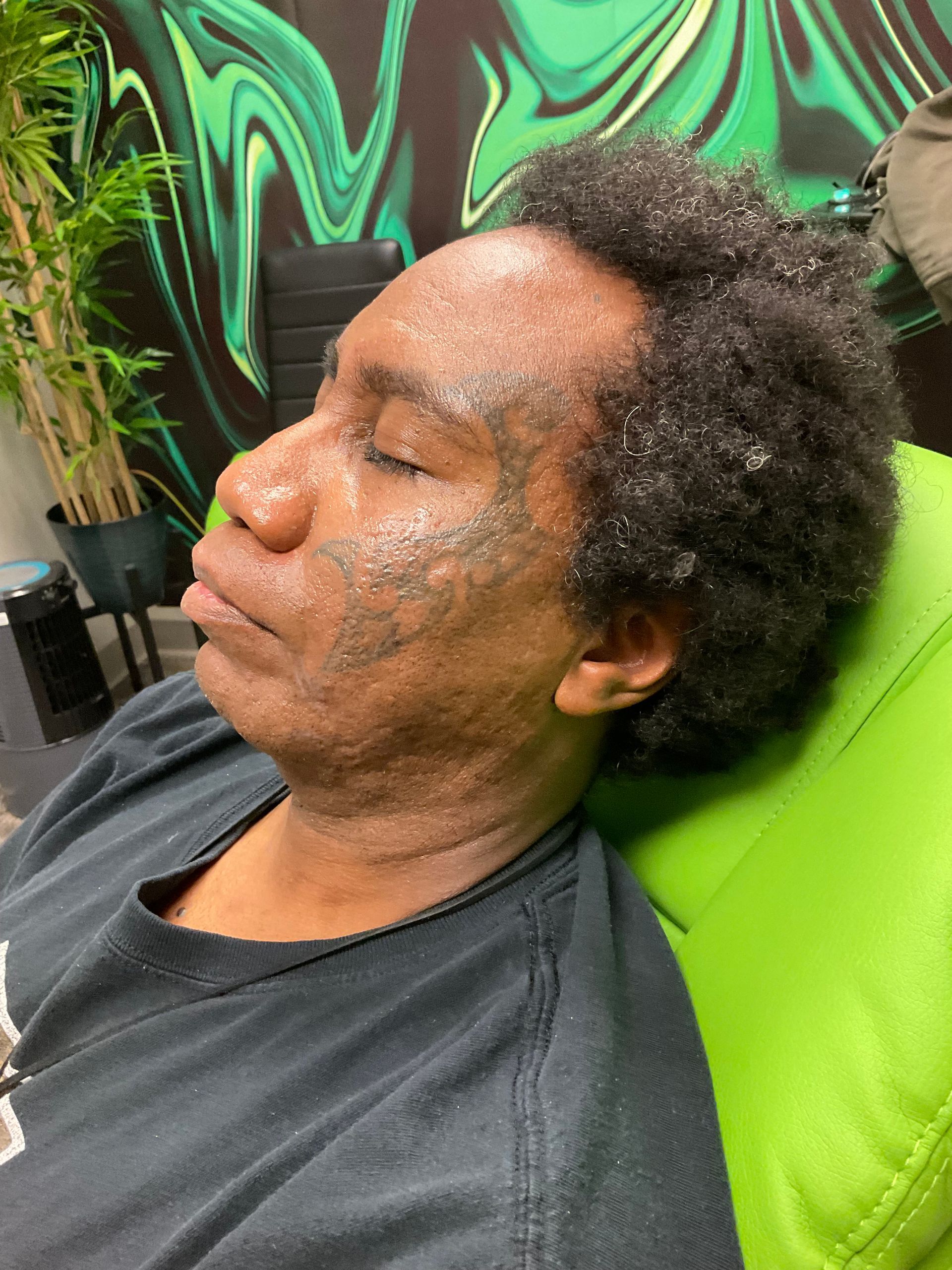 Person resting on a green chair with face markings, eyes closed, in a room with green and black wall art.