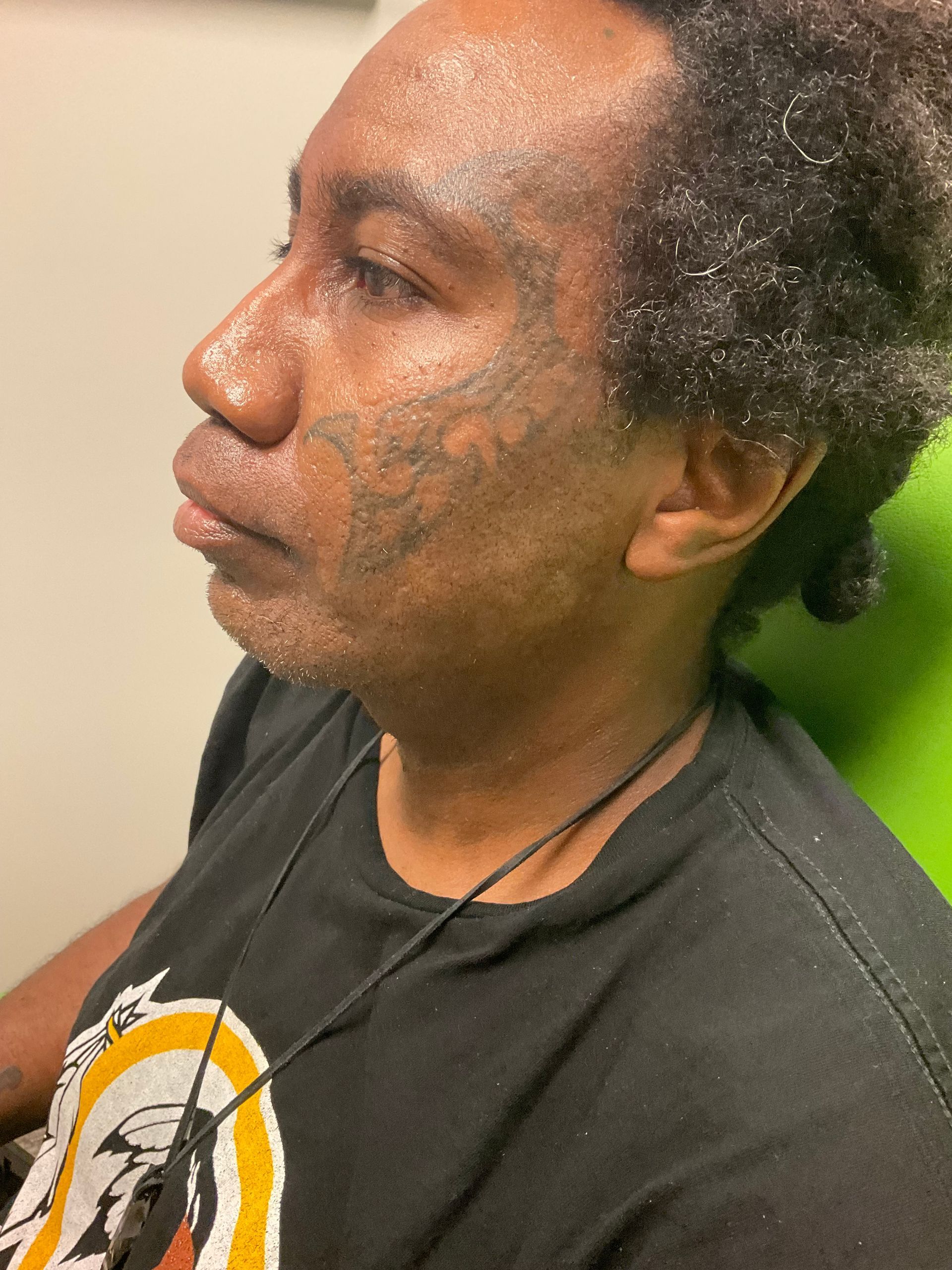 Person with face tattoos and curly hair in a black t-shirt, looking left. Sitting indoors.