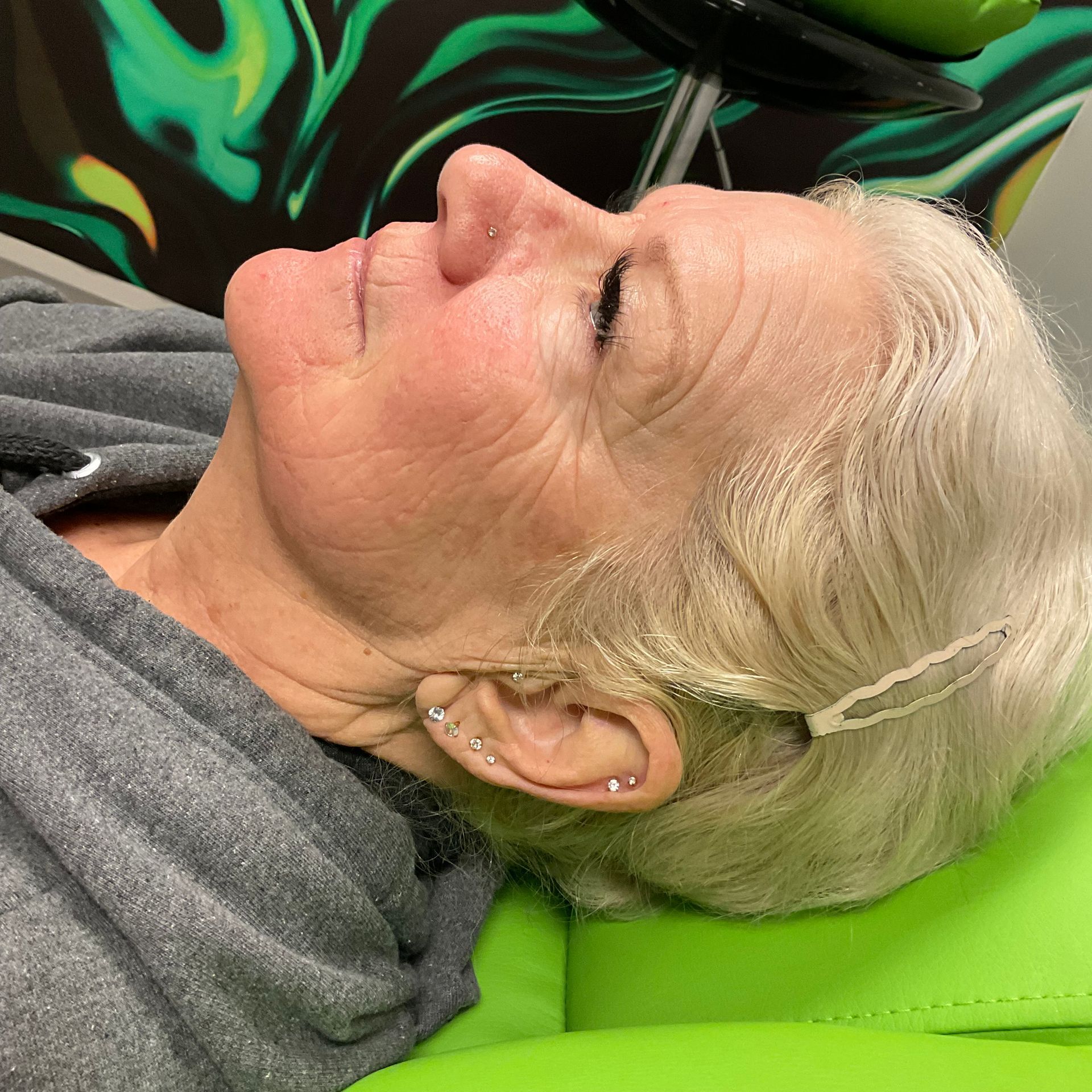 Woman lying down, side profile, gray hair with clips, green cushion, green and black background.
