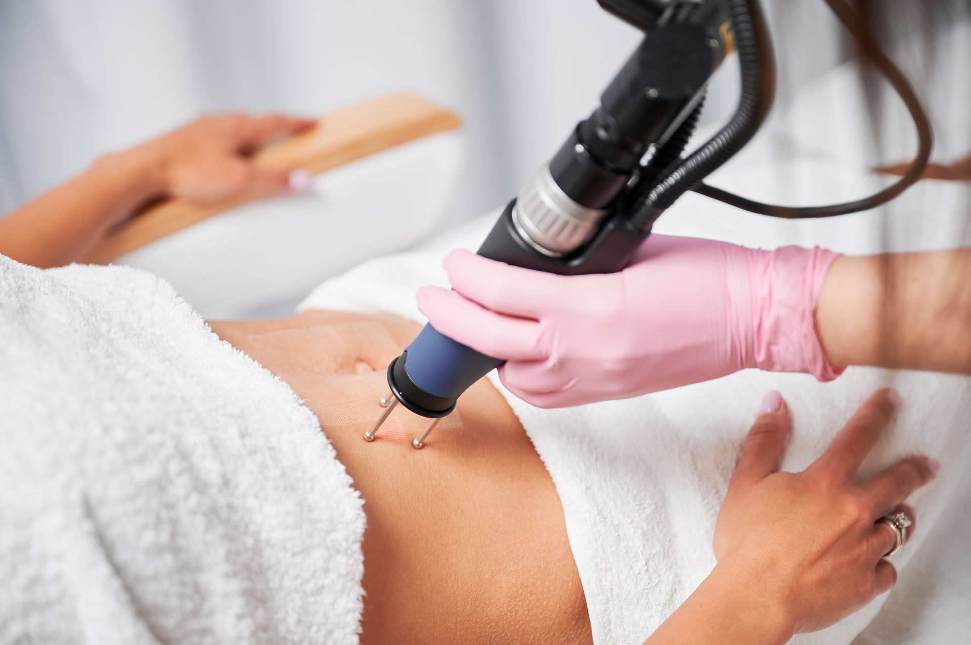 Aesthetician using a laser device on a patient's abdomen, wearing pink gloves, in a medical setting.